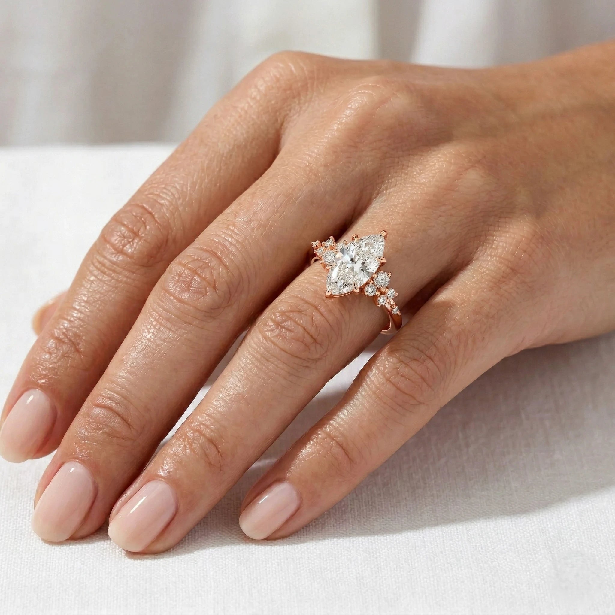 A close-up of a hand wearing a rose gold ring with a marquise-cut clear center stone and smaller side stones, resting on a white fabric surface.