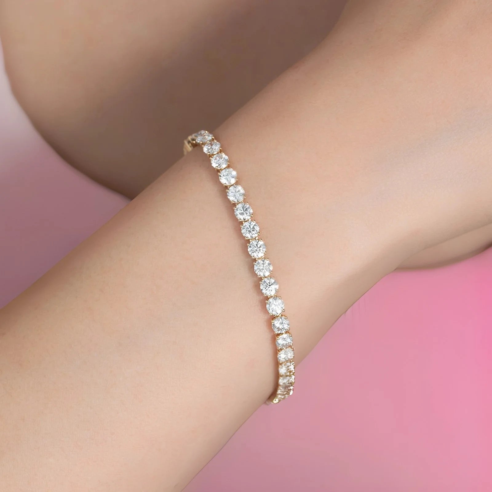 A close-up of a delicate gold tennis bracelet with round-cut clear stones worn on a person's wrist against a soft pink background.