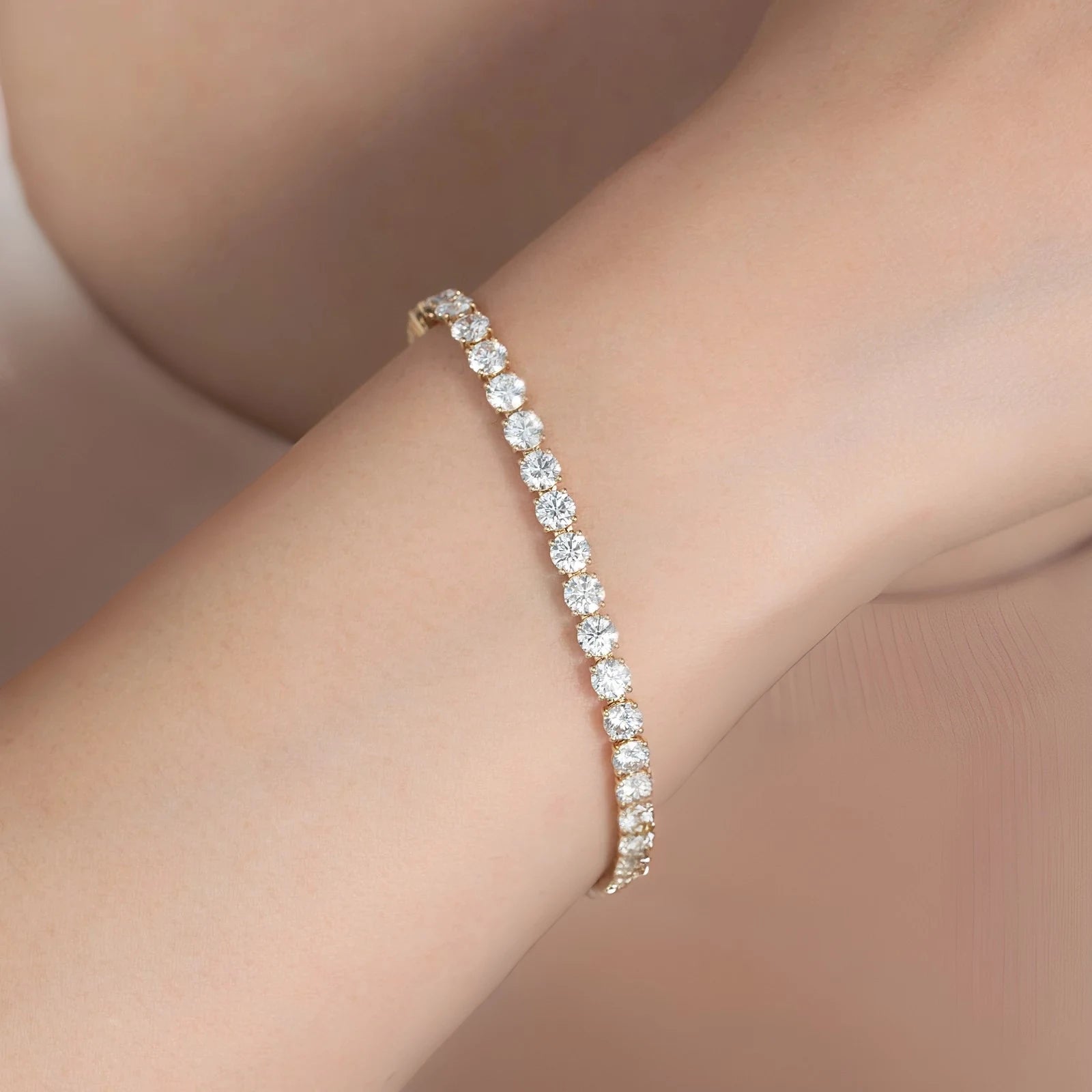 A close-up of a delicate tennis bracelet with round clear stones set in a yellow gold band, worn on a person's wrist against a soft beige background.