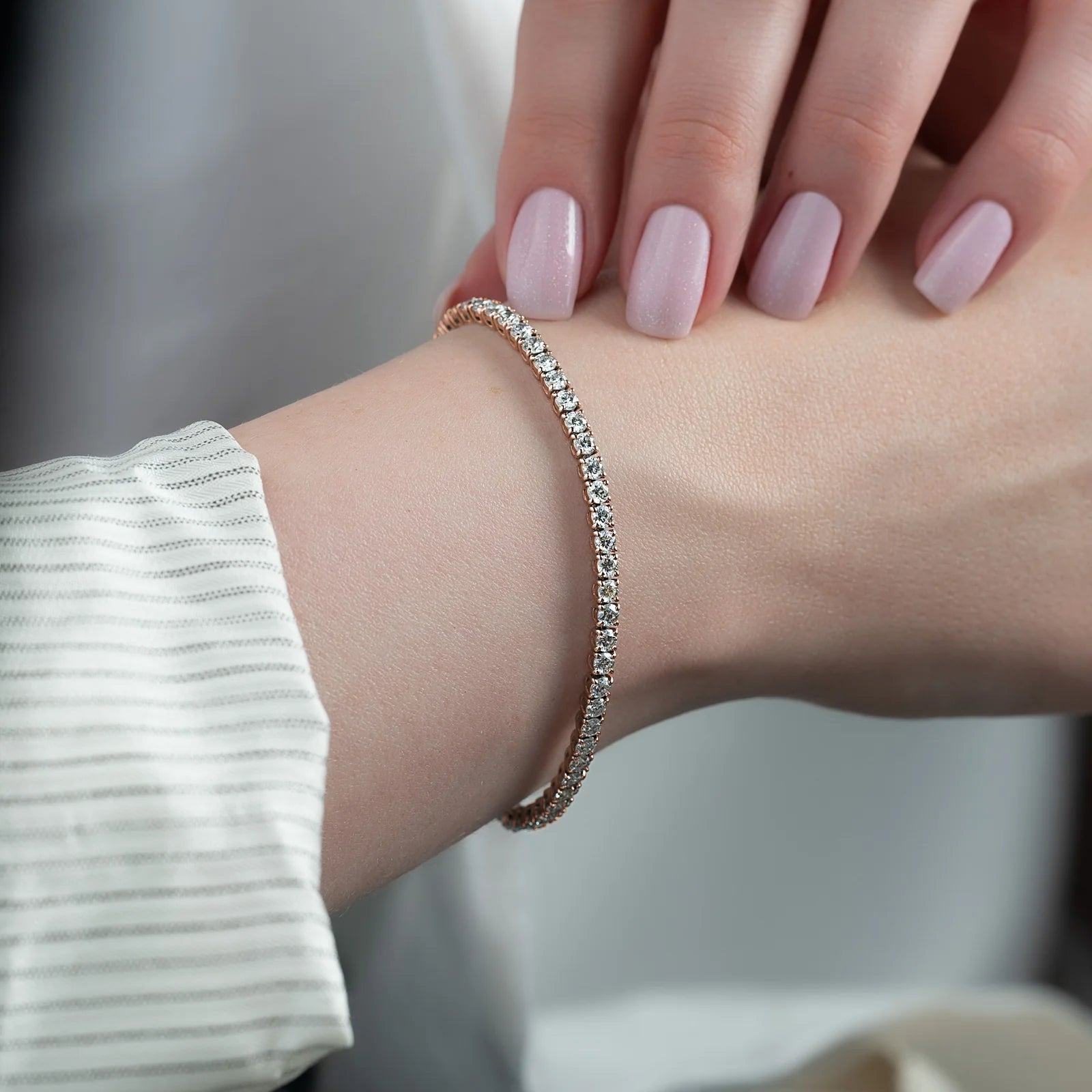 A close-up of a delicate tennis bracelet with clear stones set in a rose gold-toned band, worn on a woman's wrist. The person's hand with pale pink manicured nails rests gently above the wrist, and a white striped shirt sleeve is visible on the left.