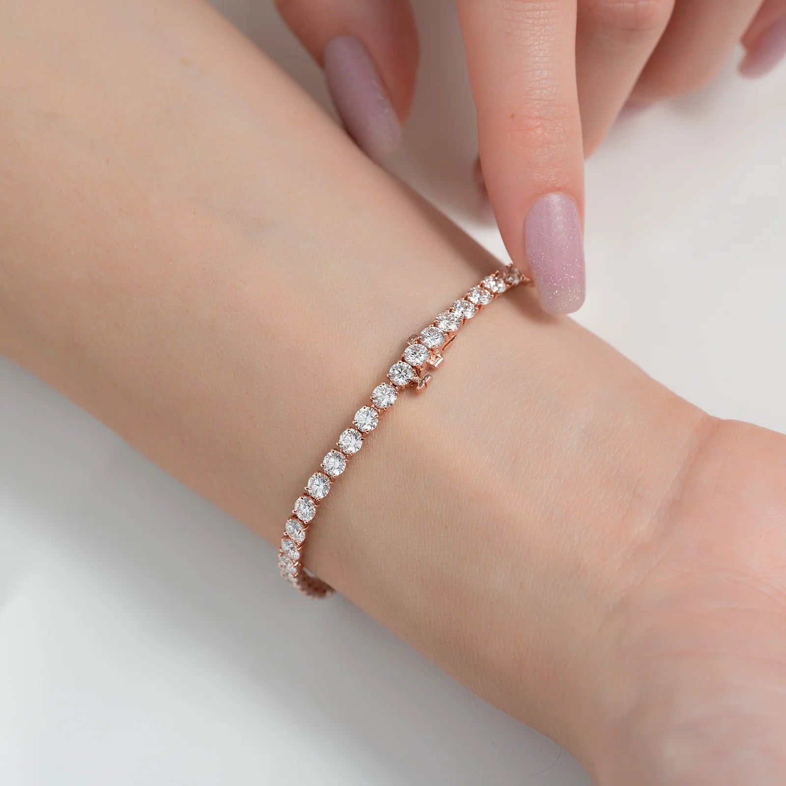 A close-up of a delicate rose gold tennis bracelet with clear round-cut stones worn on a person's wrist, with their hand resting near the top.