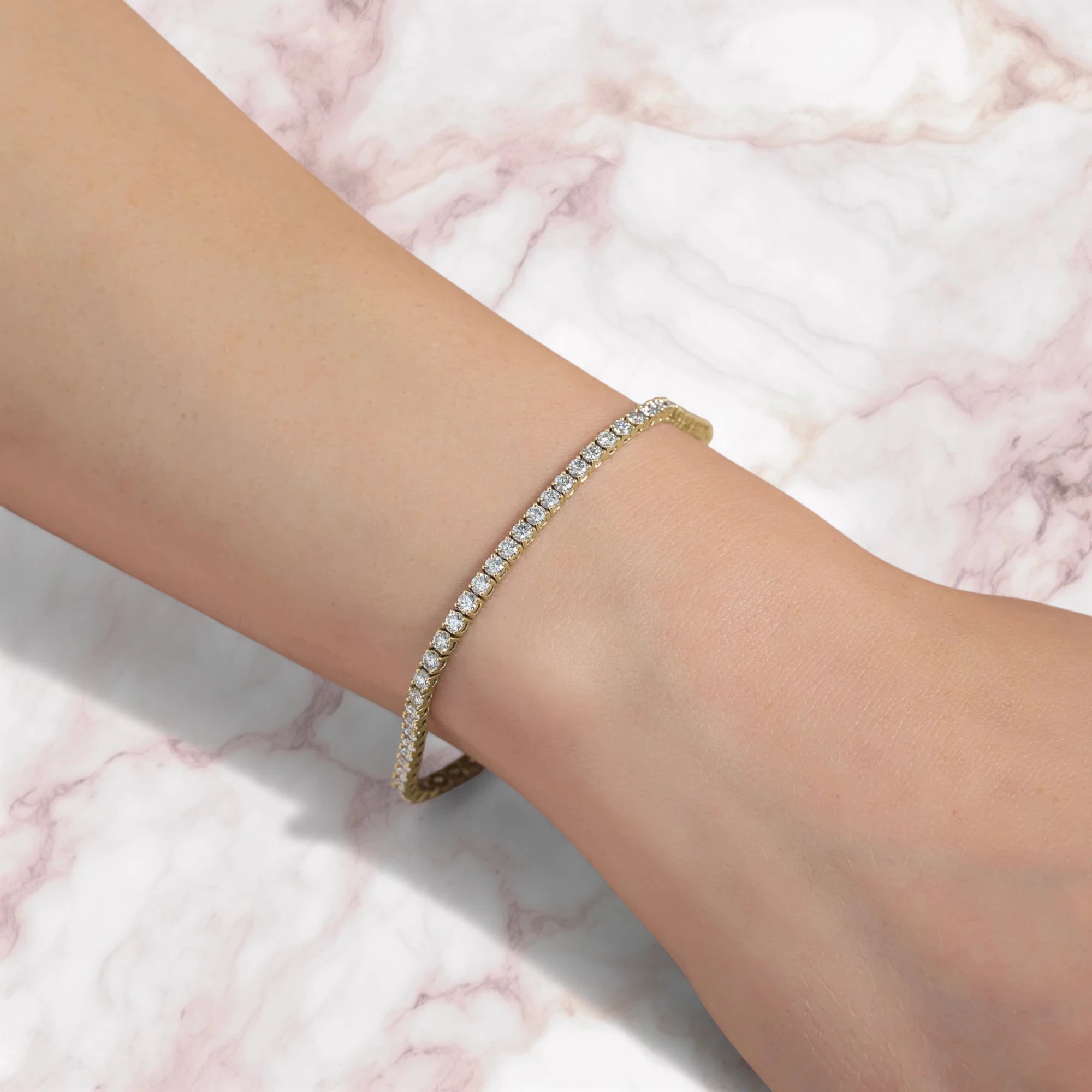 A delicate gold tennis bracelet set with small clear stones, worn on a person's wrist against a white and pink marble background.
