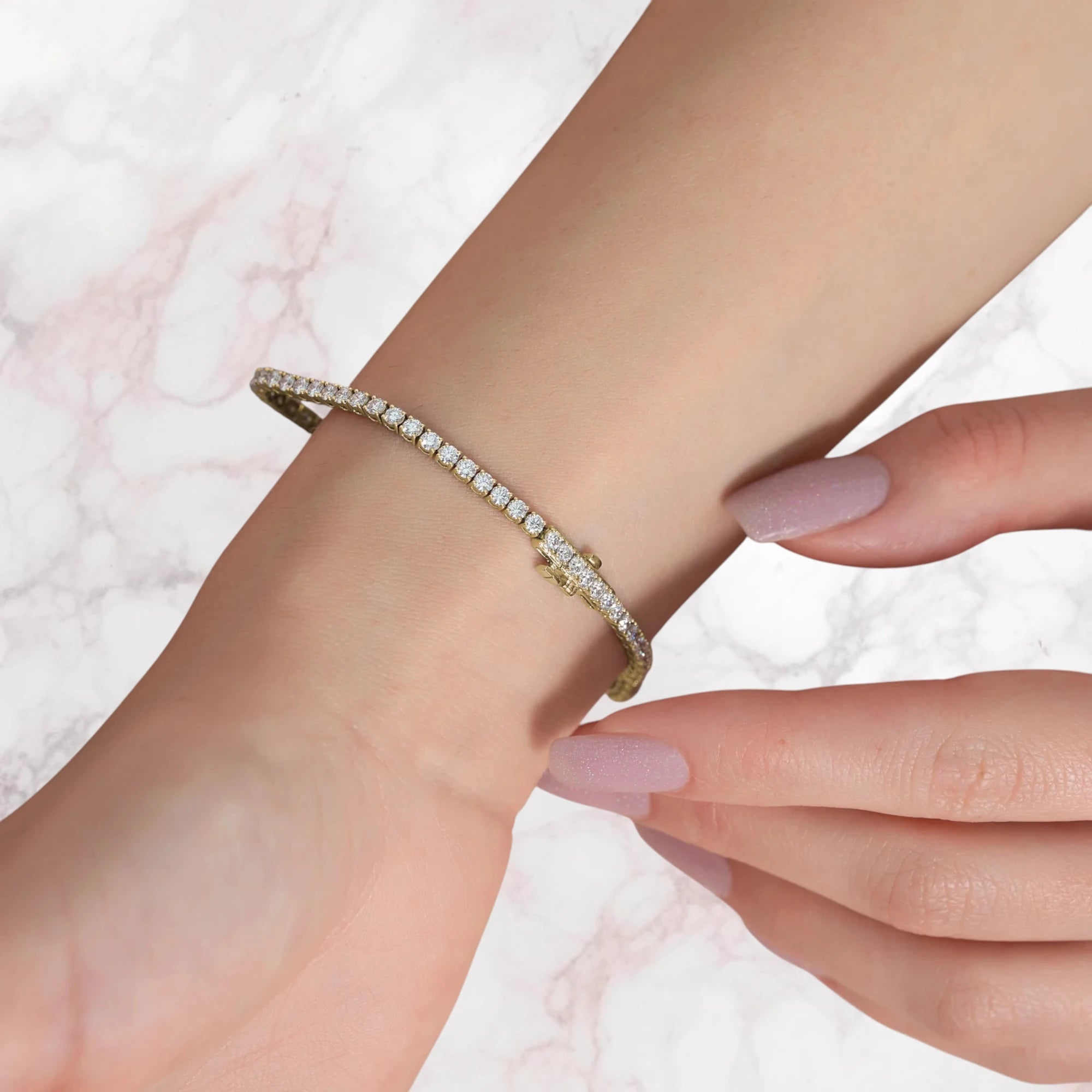 A delicate gold tennis bracelet with small clear stones, shown worn on a person's wrist against a light marble background.