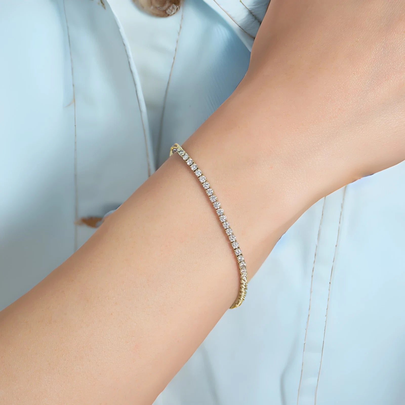 A delicate tennis bracelet with clear stones and gold-toned metal, worn on a person's wrist against a light blue shirt background.